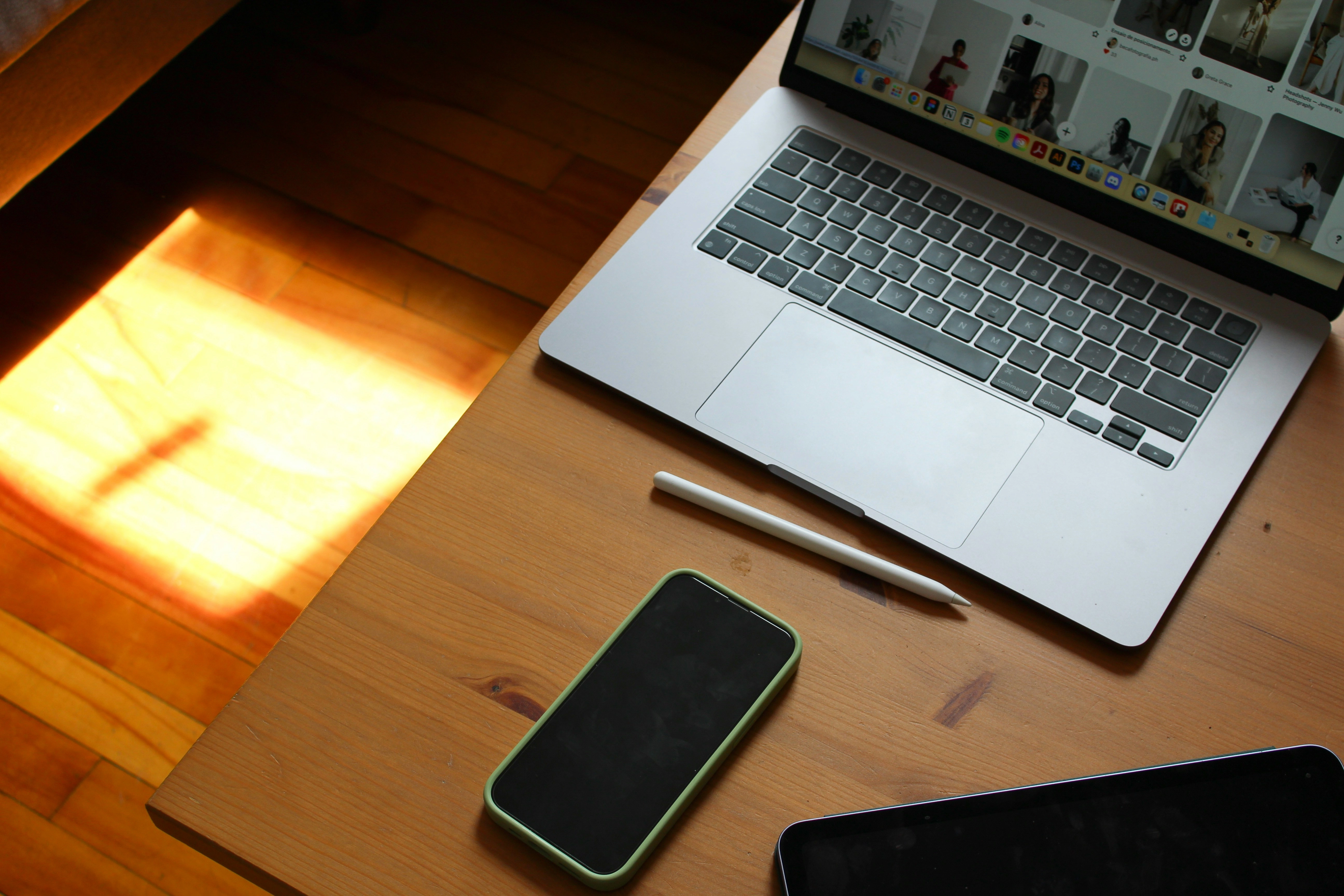 Laptop, a phone, and a pen on a wooden desk with the sun streaming in through a window.